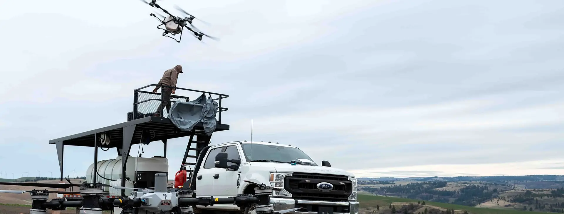 A DJI Agras spray drone lifts off above a RADS truck and platform on a gravel road with rolling hills behind