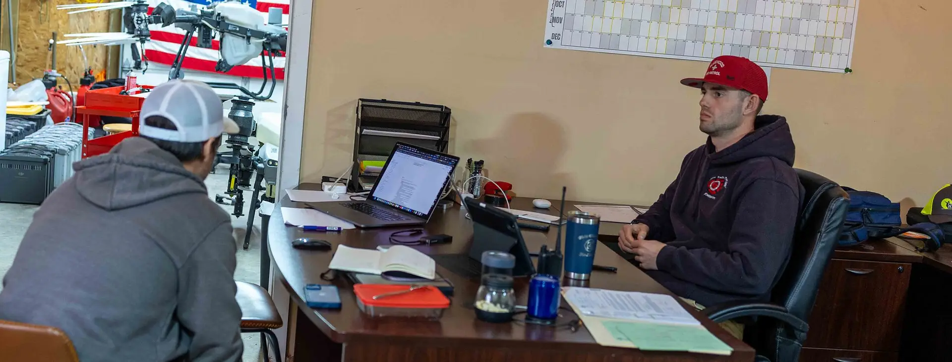 Two people talk across a desk in the RADS office with an Agras drone and parts shelving visible in the shop behind them