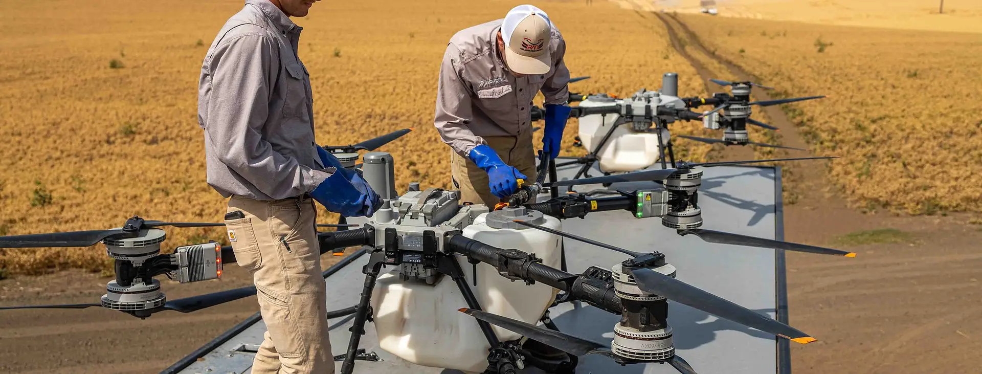 Two RADS operators in blue gloves service a DJI Agras on the platform trailer in a harvested wheat field