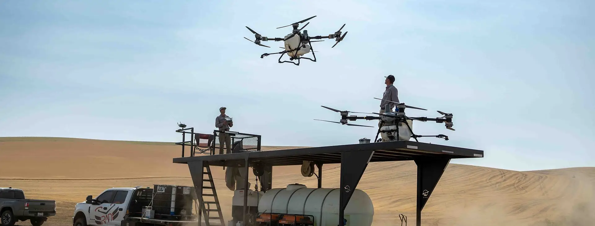 A DJI Agras drone lifts off from a RADS platform trailer in a cloud of dust at the edge of a harvested field