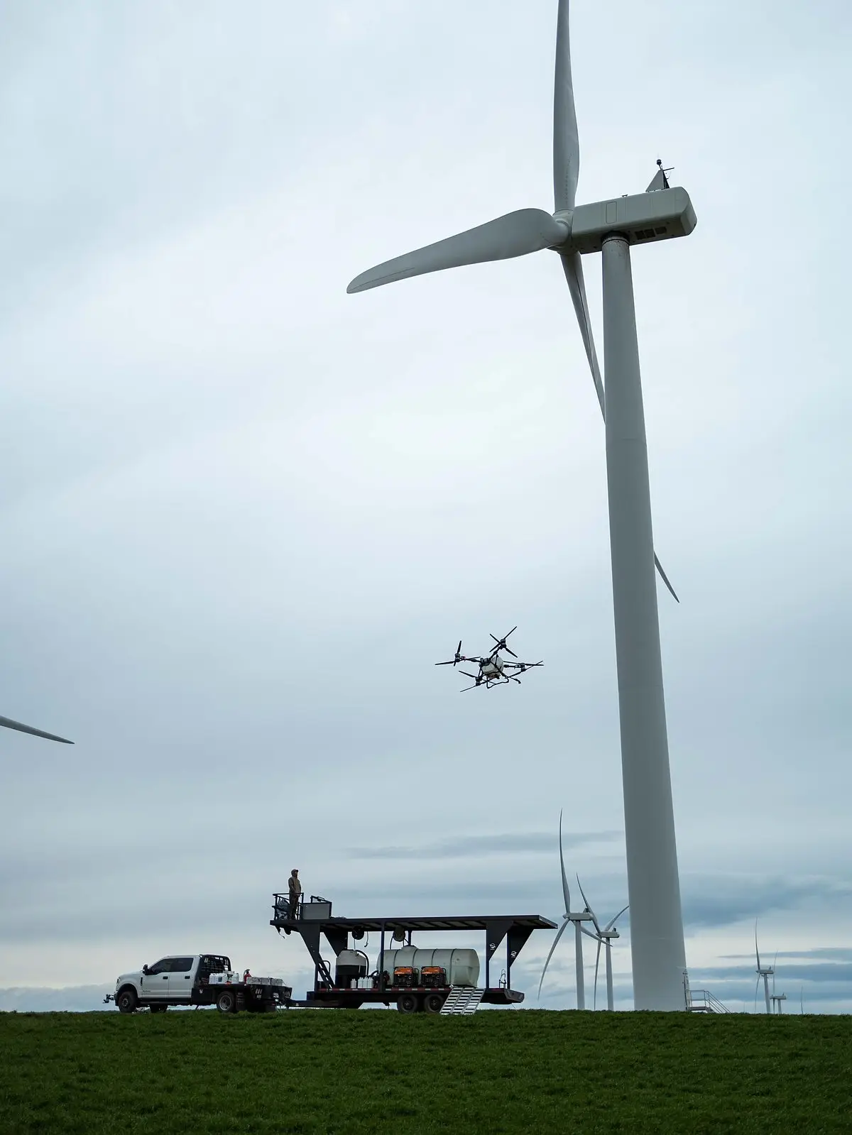 A DJI Agras drone flies beneath a wind turbine while a RADS truck and spray platform wait in a green field